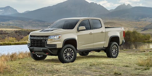 A beige colored 2022 Chevrolet Colorado truck parked on a field with a pond behind it.